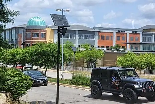 A two-way divided secondary road accessing a commercial are shown. In the foreground, which is a curbside, a green, leafy bush is shown on the bottom left, and a black pole topped with solar panel and Flock ALPR camera installed. In the midground, two vehicles can be seen driving right, a dark grey Jeep in front, a black Kia sedan behind. Behind the road and cars is a median featuring long, decorative grasses on the right side, medium-size trees on the left side. In the middle of the median, near a two-headed street lamp, another black pole topped with solar panel and Flock ALPR camera can be seen. The background shows multi-color, multi-story commercial buildings, the corner of which features a large, teal blue dome of semi-transparent material.